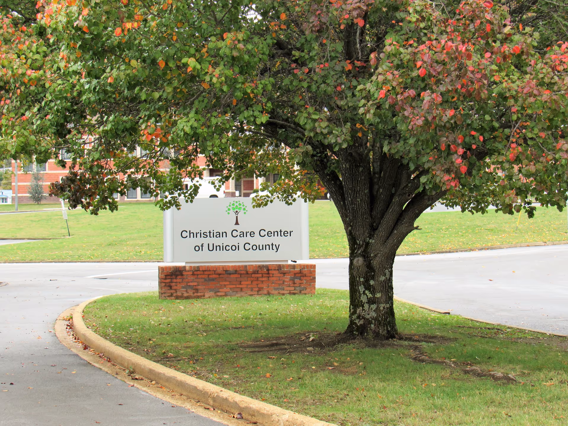 A large tree with green and red leaves stands in front of a sign that reads 'Christian Care Center of Unicoi County' on a grassy area near a curved driveway.