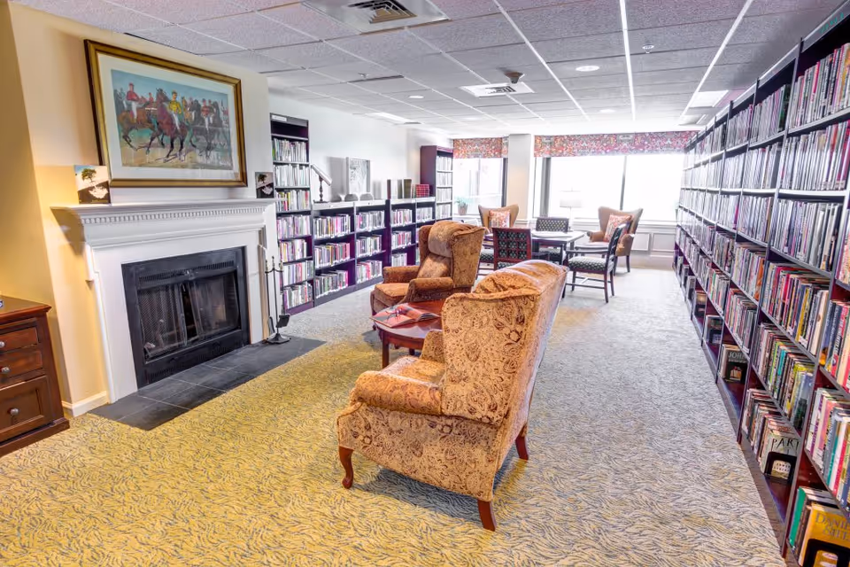 A cozy library room with patterned armchairs and a small table in the center, surrounded by tall bookshelves filled with books. There is a fireplace with a painting above it on the left side, and large windows with floral valances letting in natural light at the far end of the room.