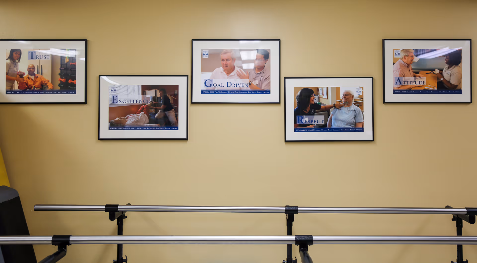 A beige wall displaying five framed motivational photos above metal parallel bars in a rehabilitation room.