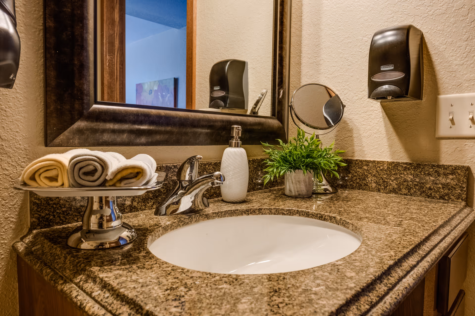 A bathroom vanity with a granite countertop, undermount sink, mirror, soap dispenser, rolled towels and a small potted plant.