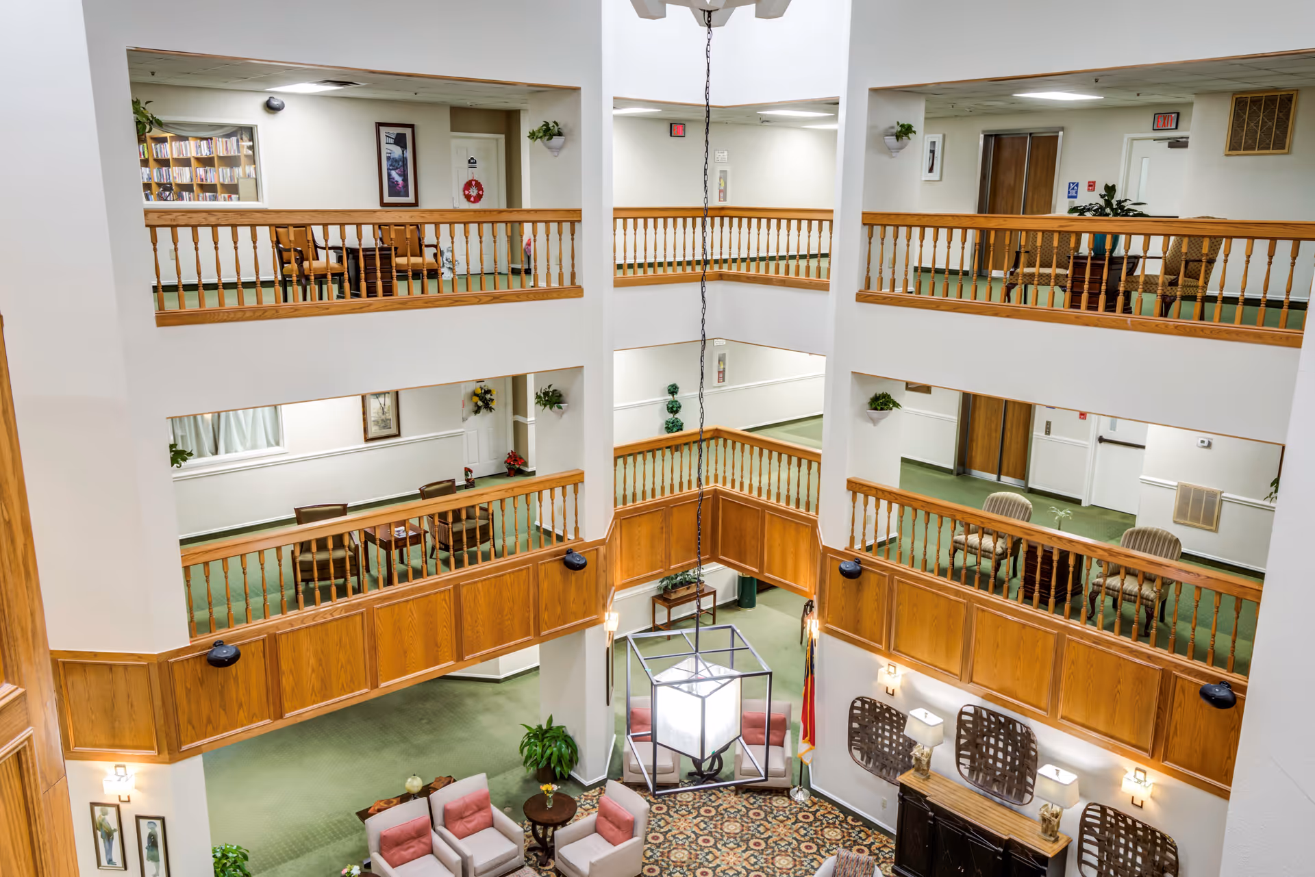 Interior view of a multi-level senior living facility common area with wooden railings, green carpeting, and seating areas with chairs and tables on each level. The lower level has a patterned carpet and a seating arrangement with armchairs and a small table. The walls are white with some framed pictures and plants placed around.
