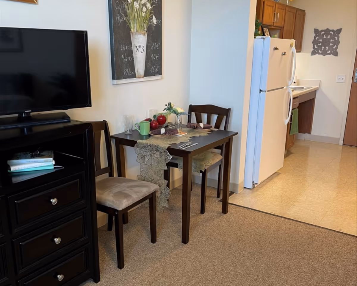 A small dining area with a dark wooden table set for two, featuring a lace table runner, two place settings with napkins, and a small flower vase. Two wooden chairs with cushioned seats are placed at the table. To the left, there is a black dresser with a flat-screen TV on top and some books in an open shelf. In the background, a kitchen area with a white refrigerator, wooden cabinets, and a sink is visible. The floor transitions from carpet in the dining area to linoleum in the kitchen. A decorative wall art piece hangs above the dining table.