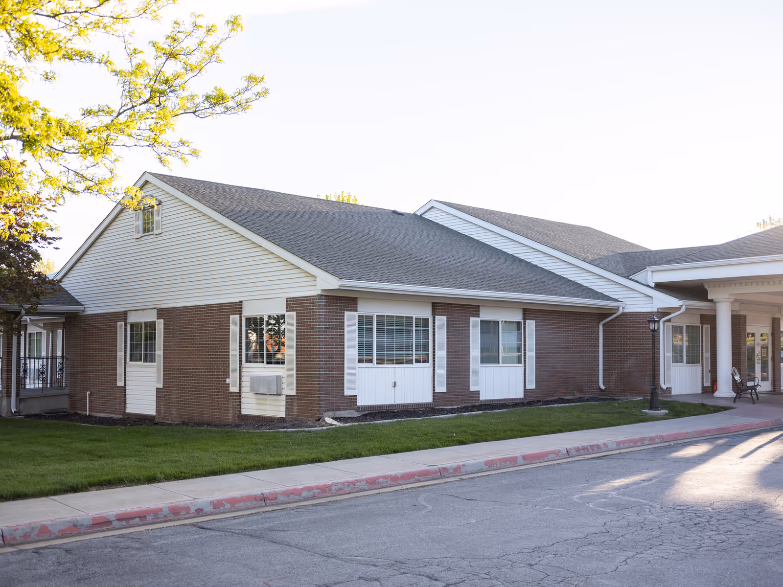 Exterior view of a single-story brick and siding building with multiple windows, a covered entrance supported by white columns, a sidewalk, and a small grassy area with trees.