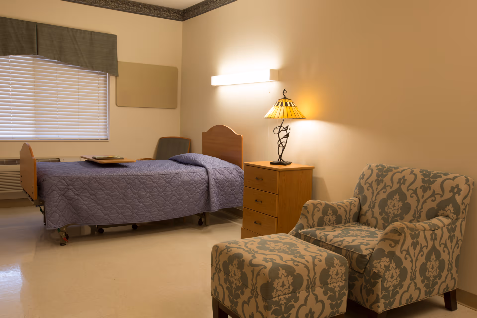 A senior resident room with a single bed draped in a purple quilt, a wooden nightstand and lamp, and a patterned armchair with ottoman.