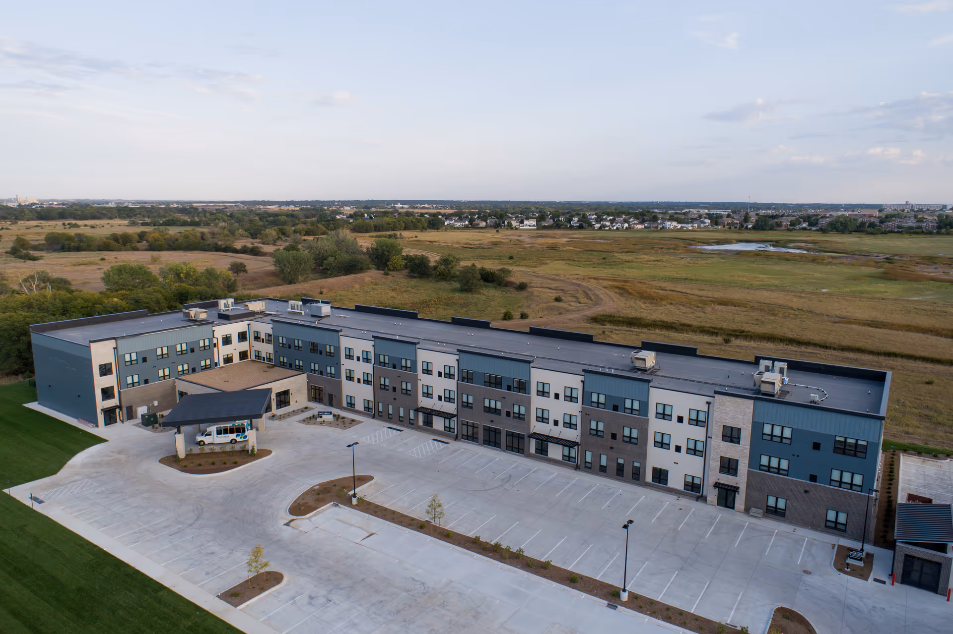 Aerial view of a modern three-story senior living facility building surrounded by open fields and a large empty parking lot under a partly cloudy sky.
