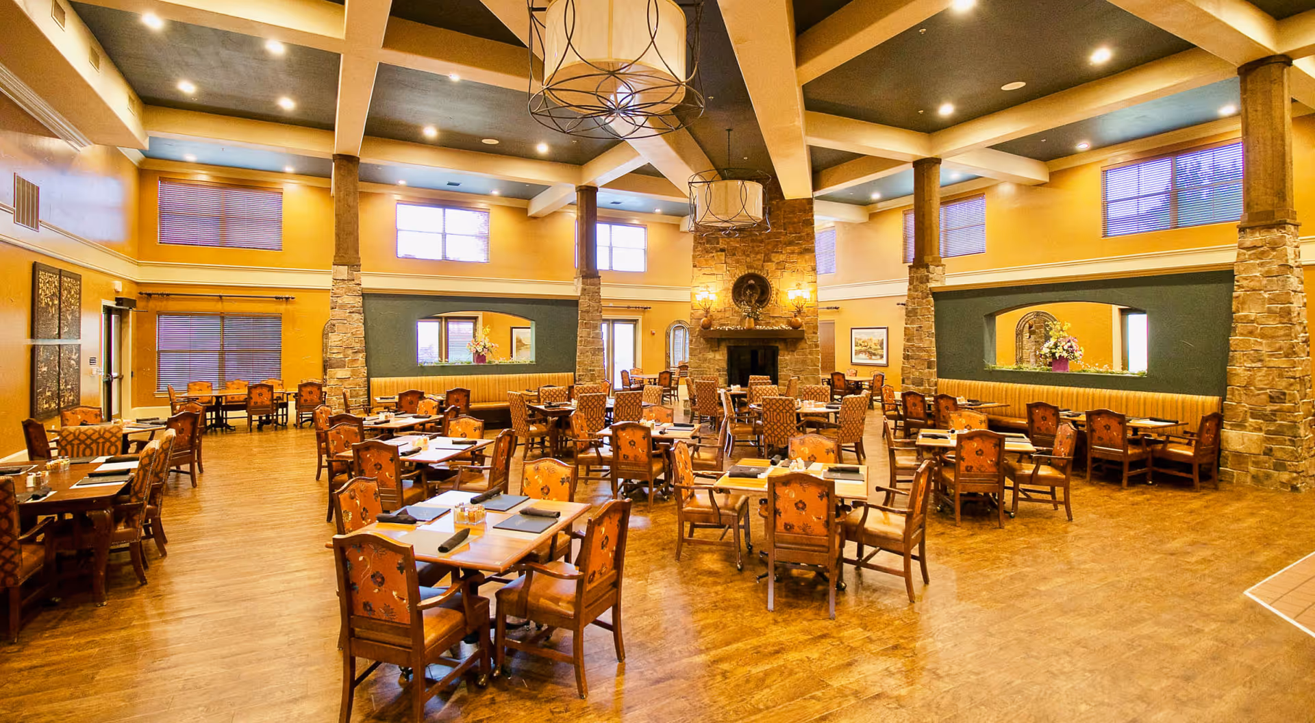 A spacious dining room with multiple wooden tables and chairs arranged neatly. The room features high ceilings with exposed beams, large windows with blinds, stone pillars, and a stone fireplace in the center. Warm lighting and decorative elements create a cozy and inviting atmosphere.