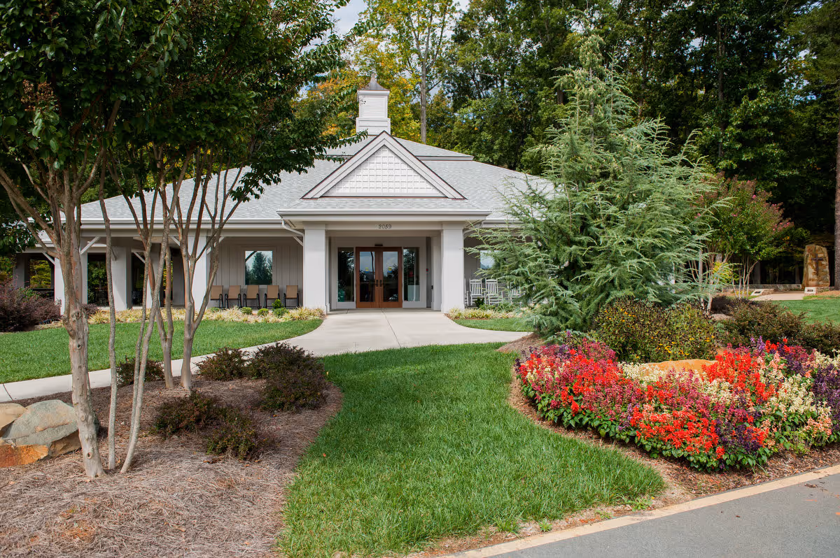 Front exterior view of a single-story building with a gray roof and white walls, surrounded by well-maintained landscaping including green grass, trees, and colorful flower beds. A concrete walkway leads to the double glass entrance doors.