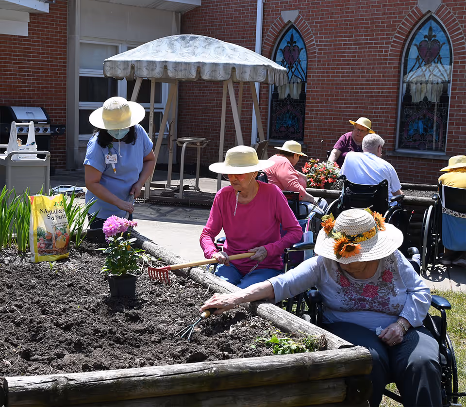 Several elderly individuals, some in wheelchairs, wearing sun hats and gardening outdoors in a raised garden bed. A caregiver wearing a mask and hat is assisting with the gardening. The background shows a brick building with stained glass windows and a covered swing.