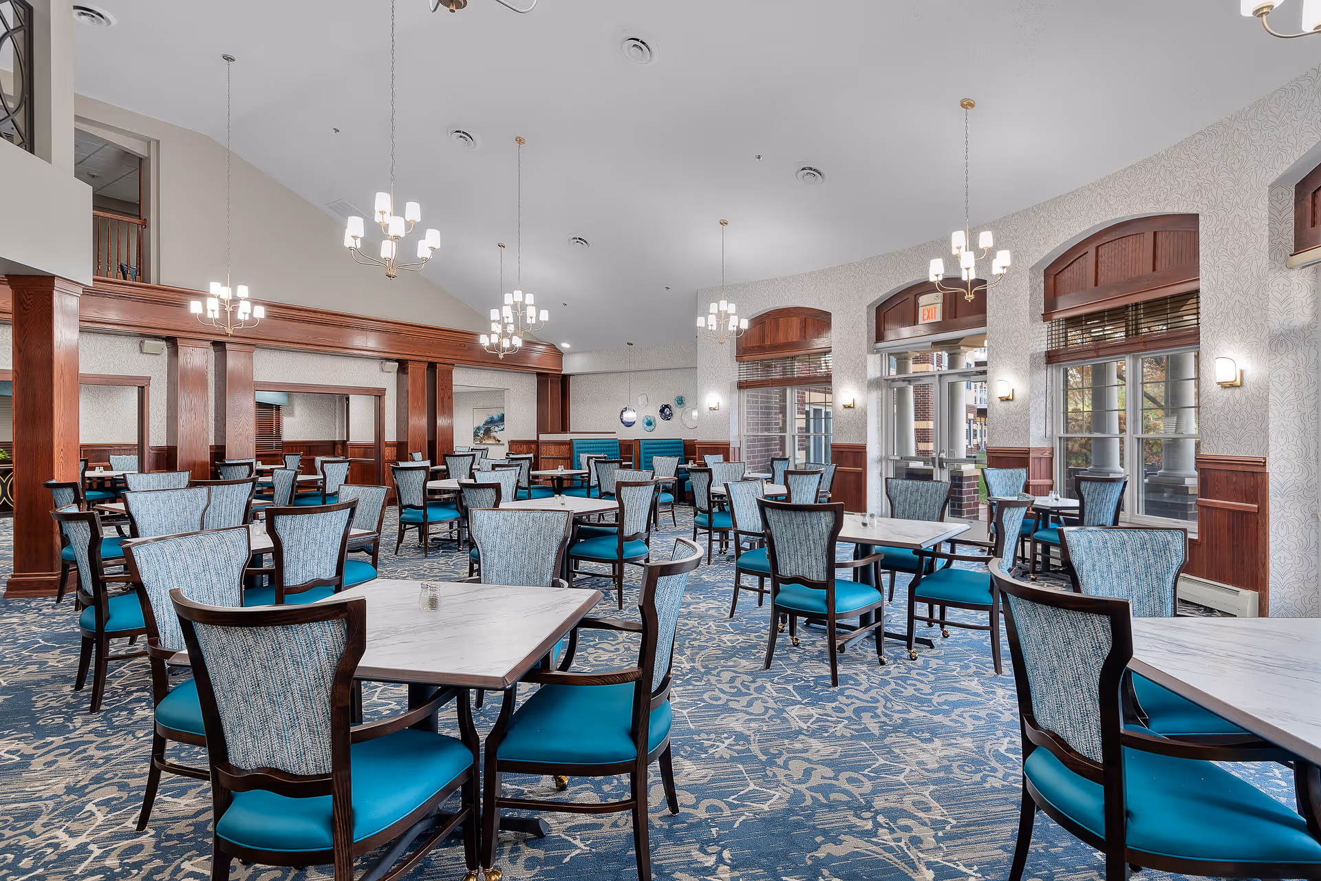 A spacious dining room with multiple tables and chairs arranged neatly. The chairs have blue cushions and patterned backs, and the tables have white marble tops. The room features large windows with wooden frames, allowing natural light to enter. Elegant chandeliers hang from the high ceiling, and the walls are decorated with wood paneling and light-colored wallpaper. The carpet has a blue and beige floral pattern.