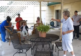 A group of elderly people and caregivers are gathered in a covered outdoor patio area. One elderly woman is watering a potted plant on a round metal table with chairs around it. Other individuals are seated or standing nearby, engaging with each other.