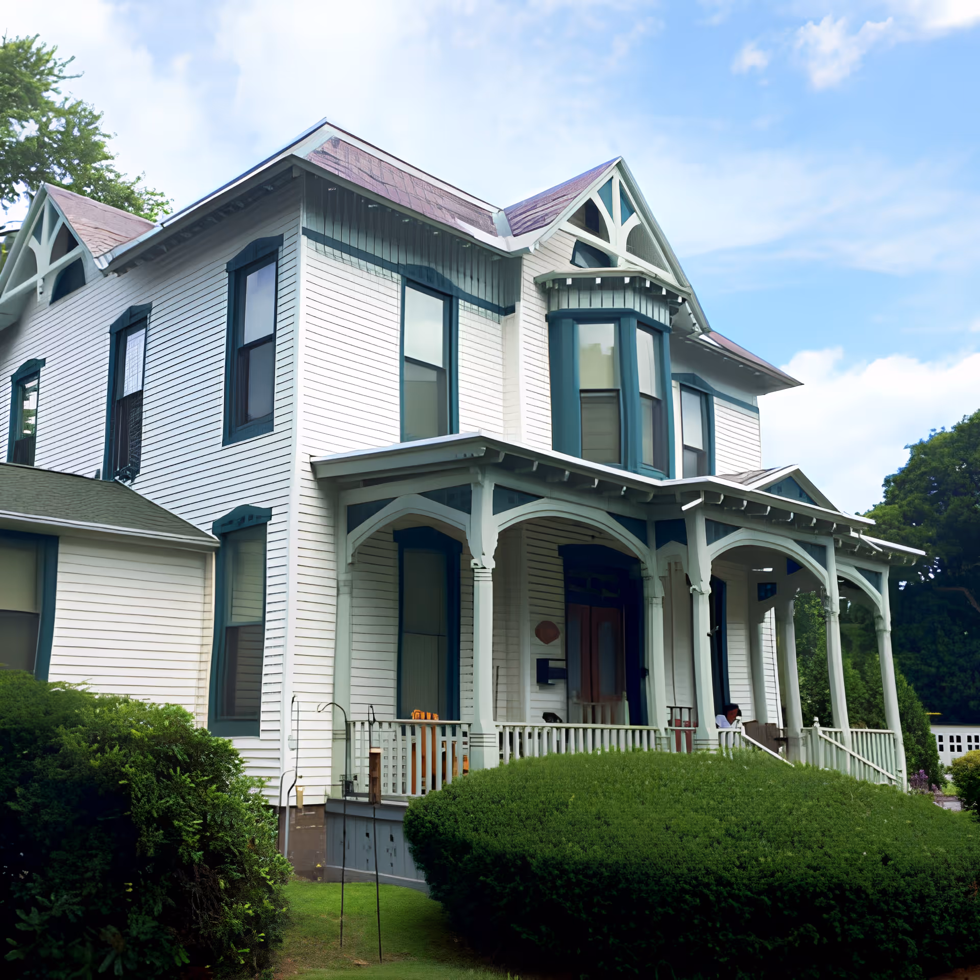 A large, two-story white house with green trim and a covered front porch surrounded by well-maintained bushes and greenery under a partly cloudy blue sky.