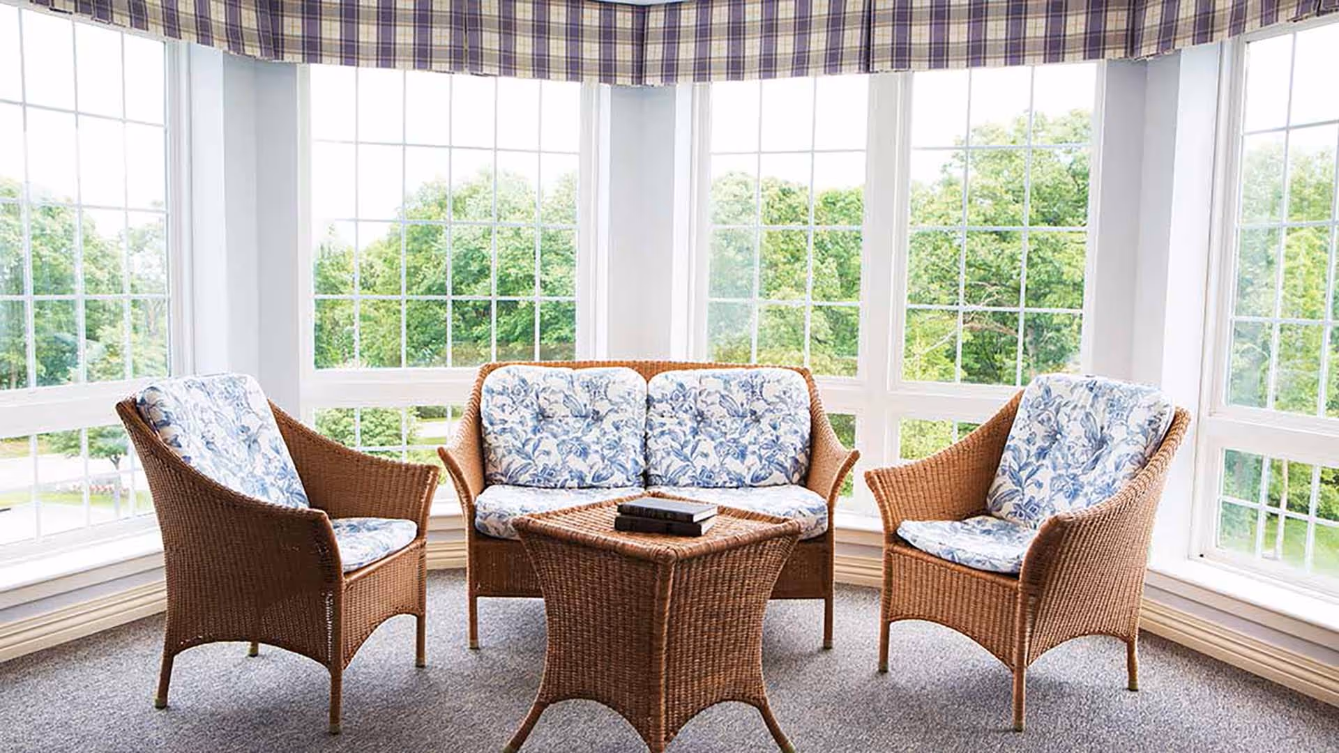 A bright sunroom with large windows showing green trees outside. The room contains wicker furniture including a loveseat and two armchairs with blue and white floral cushions, and a matching wicker coffee table with books on it. The windows have a checkered valance at the top.