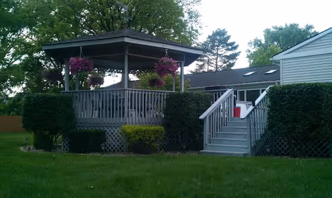 Backyard deck with a covered gazebo, hanging flower baskets, stairs and lawn in front of a single-story building.