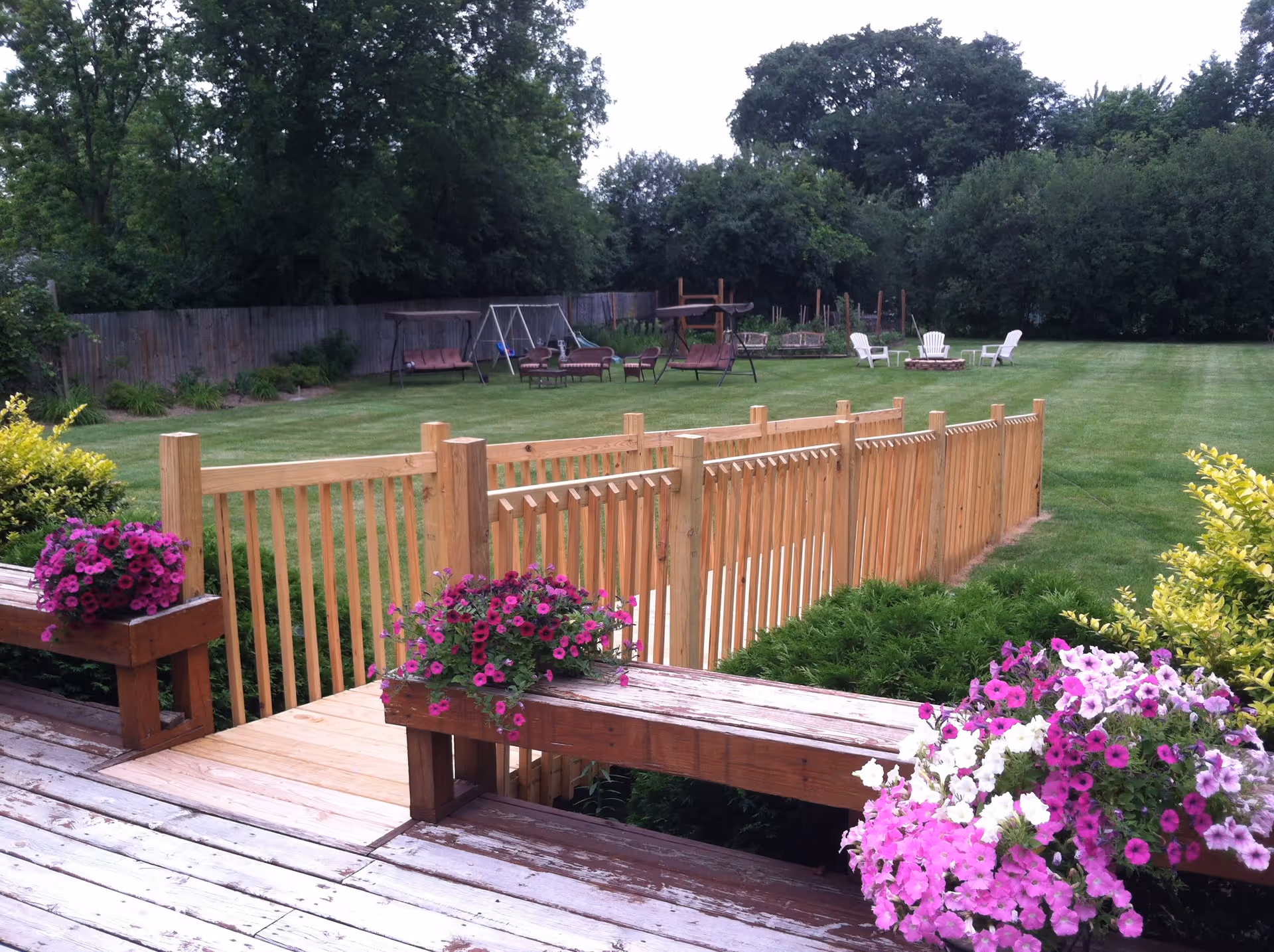 A spacious outdoor garden area with a wooden deck in the foreground adorned with vibrant pink and white flowers in planters. A wooden fence runs through the middle of the grassy lawn. In the background, there are several outdoor seating options including swings, chairs, and a fire pit surrounded by white chairs. Trees and bushes border the area.
