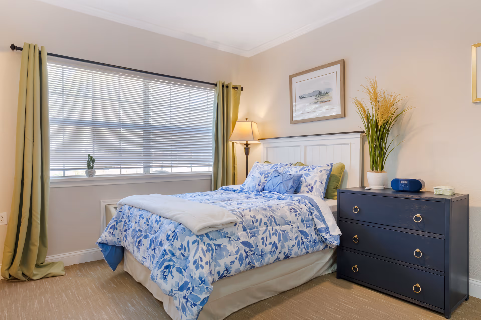 A bright bedroom with a bed featuring blue and white floral bedding, a white headboard, and green pillows. Next to the bed is a dark blue dresser with three drawers, topped with a potted plant, a blue radio, and a tissue box. A floor lamp stands in the corner near a window with closed blinds and green curtains. A framed picture hangs on the beige wall above the headboard.
