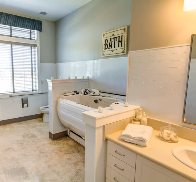 A bright accessible bathroom featuring a walk-in bathtub, a sink vanity with folded towels and toiletries, and a toilet by a window.