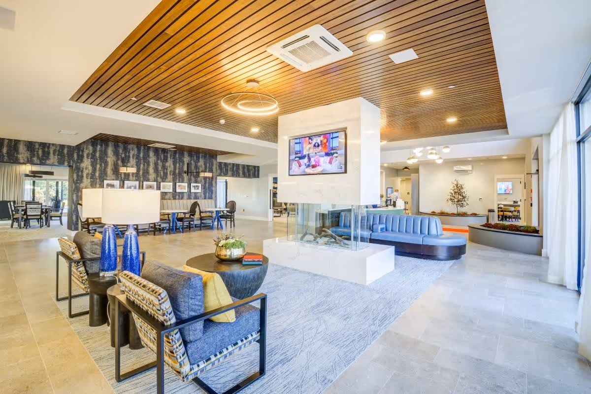 A spacious and modern living room area in a senior living facility featuring a central white marble fireplace with a mounted TV above it. The room has comfortable seating including blue cushioned chairs, patterned armchairs with yellow pillows, and a round coffee table with decorative items. The ceiling has wooden slats with recessed lighting and a circular pendant light. Large windows with sheer curtains allow natural light to fill the space. In the background, there are additional seating areas and a dining area with tables and chairs.