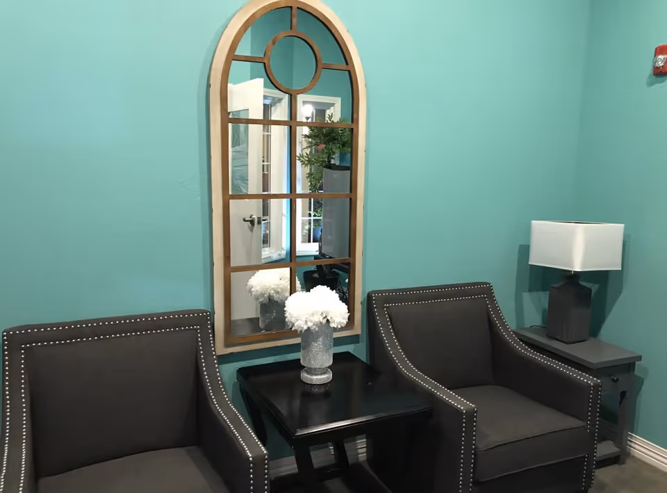 A cozy seating area with two dark gray armchairs featuring silver nailhead trim, separated by a black side table with a vase of white flowers. A decorative arched mirror with a wooden frame hangs on a teal wall behind the chairs. To the right, there is a gray side table with a modern white lampshade on a dark base.