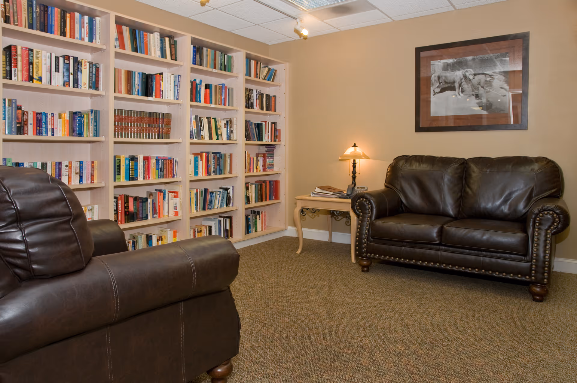 A cozy room with beige walls and carpeted floor featuring a dark brown leather loveseat and matching armchair. There is a wooden side table with a lamp and some books on it. The back wall has built-in bookshelves filled with various books, and a framed black and white photograph hangs above the loveseat.
