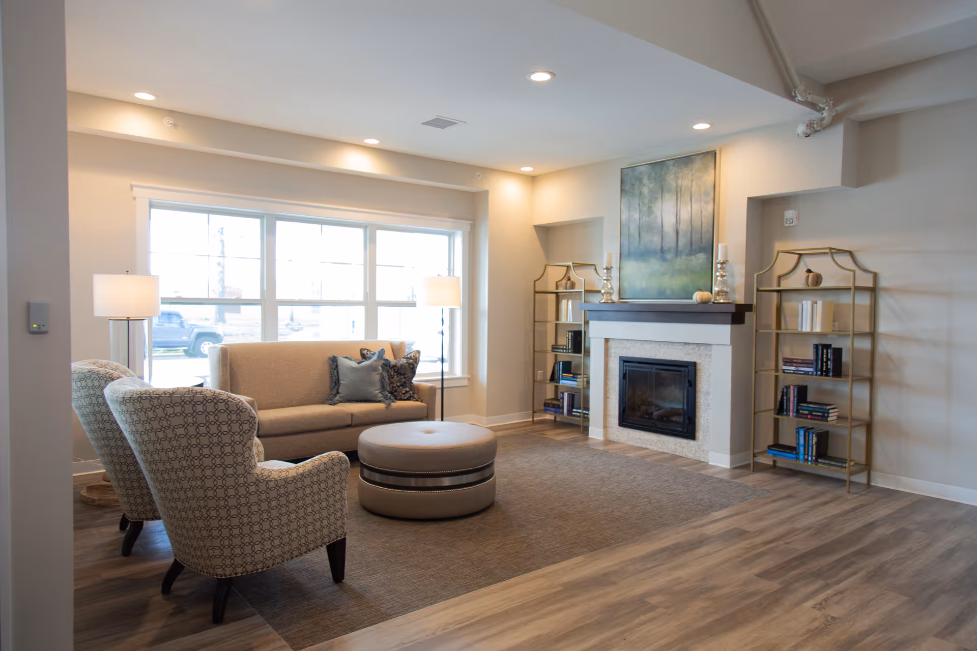 A cozy living room area with a beige sofa, two patterned armchairs, a round ottoman, and two floor lamps. There is a fireplace with a large painting above it and two gold metal bookshelves on either side holding books and decorative items. The room has large windows letting in natural light and wood flooring.