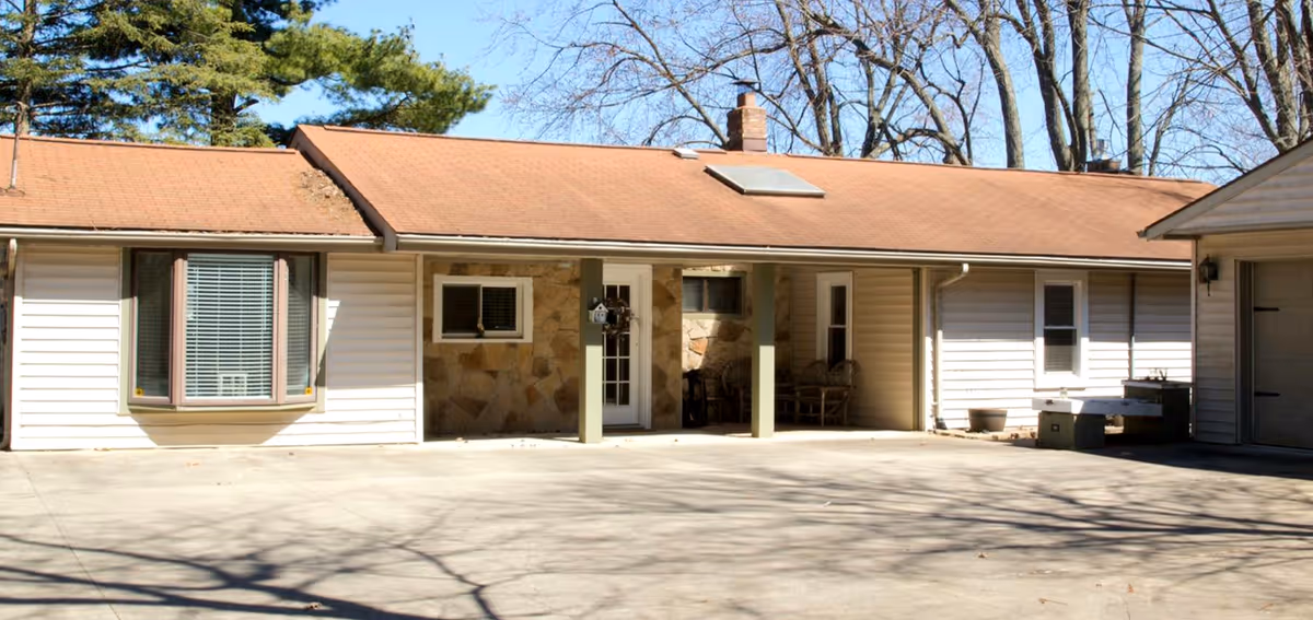Single-story building with a brown shingled roof and white siding, featuring a covered porch area with two green pillars and a stone wall section around the entrance door. There is a large concrete driveway in front and some leafless trees in the background under a clear blue sky.