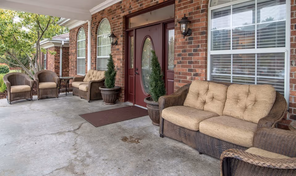 Outdoor covered patio area with wicker furniture including a cushioned sofa, two armchairs, and a small table. The patio is adjacent to a brick building with large windows and a maroon door with an oval glass panel. Two potted plants flank the door.