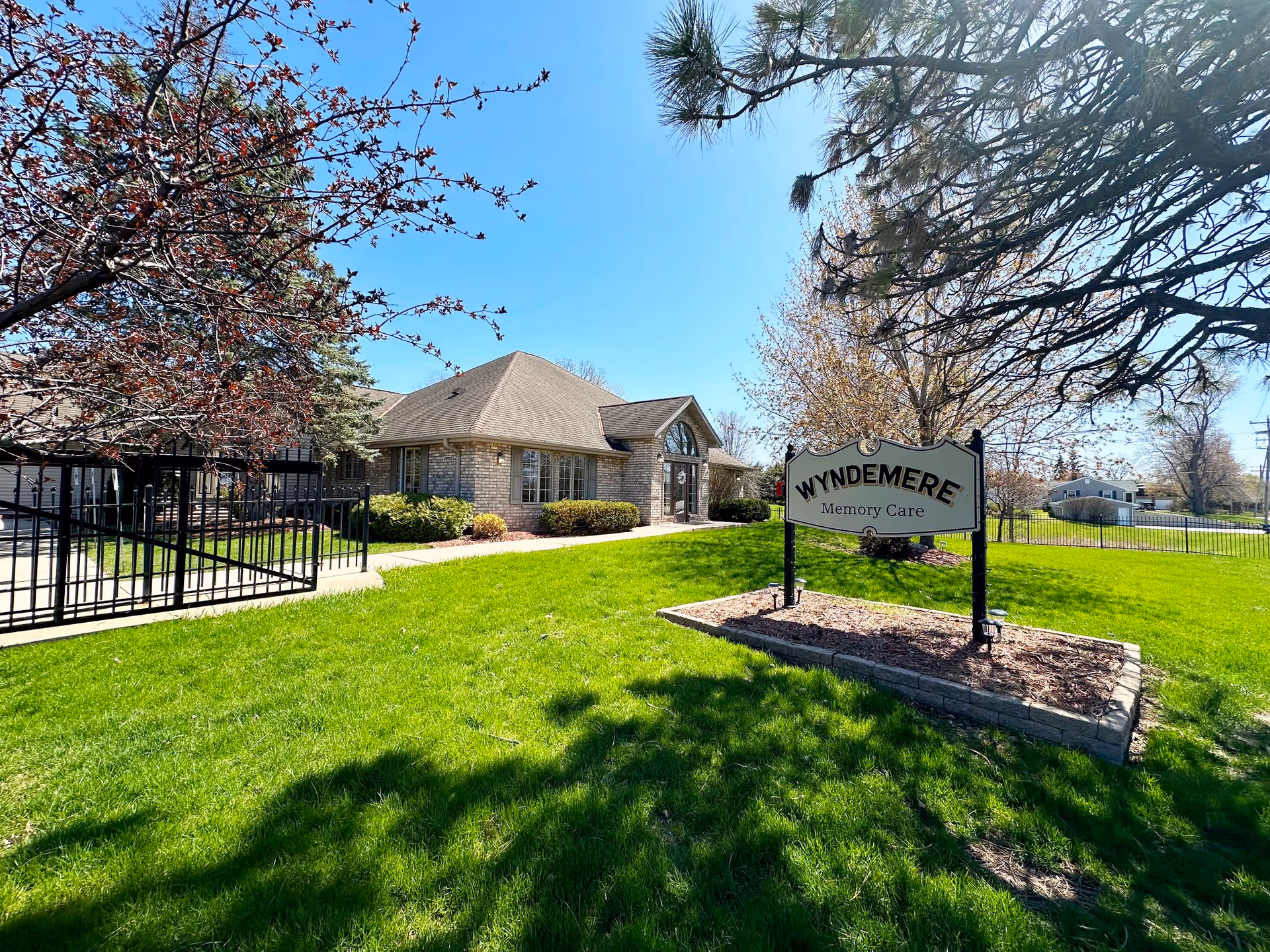 Exterior view of Wyndemere Memory Care facility showing a single-story brick building with a gabled roof, surrounded by green grass, trees with budding leaves, and a black metal fence. A sign in the foreground reads 'Wyndemere Memory Care'.