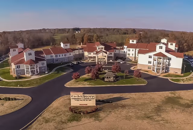 Aerial view of Fieldstone Place, a senior living facility with multiple connected buildings featuring red roofs and white walls. The facility is surrounded by paved roads, landscaped areas with trees, and open fields in the background. A sign at the entrance displays the name and contact information of the facility.