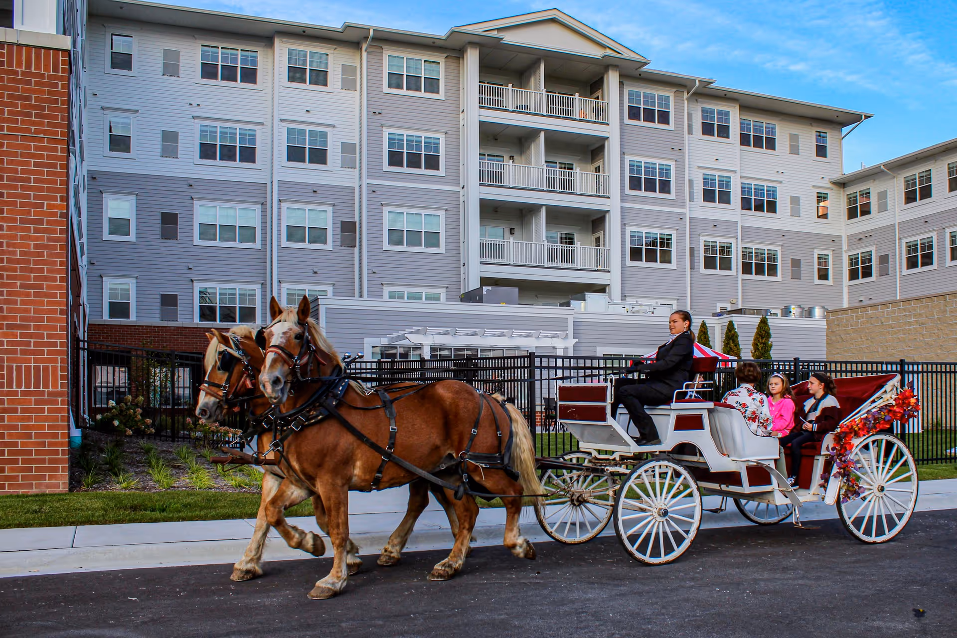 A horse-drawn white carriage carrying passengers passes in front of a multi-story senior living building.