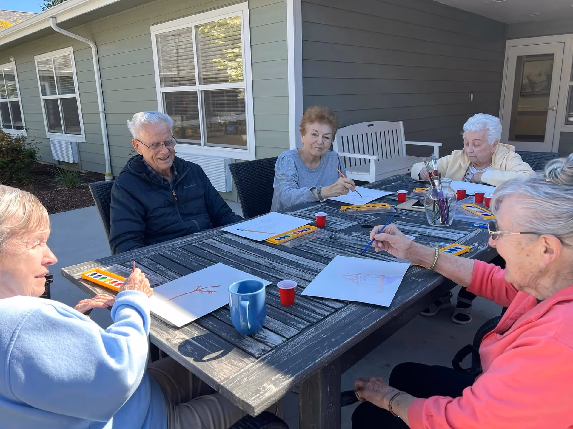 Five elderly individuals sitting around a wooden outdoor table engaged in painting activities with watercolor sets and brushes. They are outside a building with green siding and white trim, enjoying a sunny day.