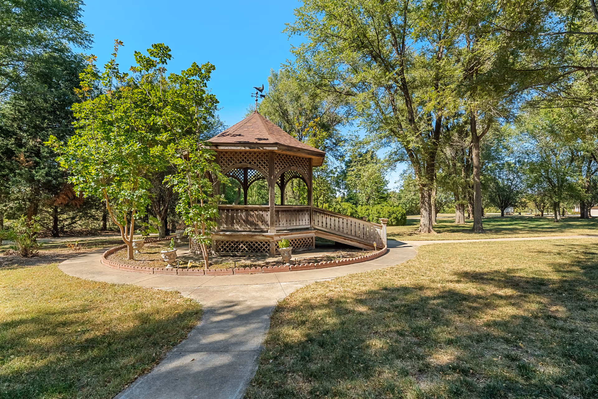 A wooden gazebo with a shingled roof and ramp access is situated in a grassy park area surrounded by trees under a clear blue sky.