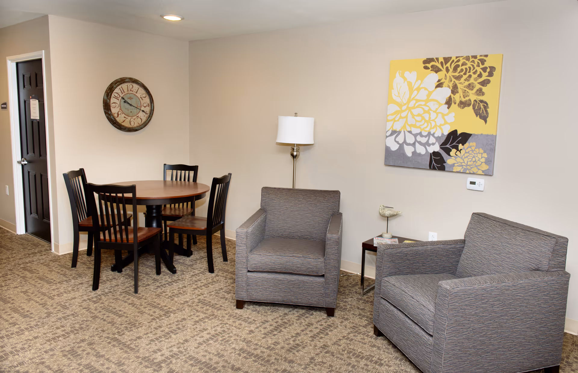 A cozy interior room with two gray armchairs, a small side table with a decorative bird, a floor lamp, a round wooden dining table with four chairs, a wall clock, and a large floral artwork on the wall.