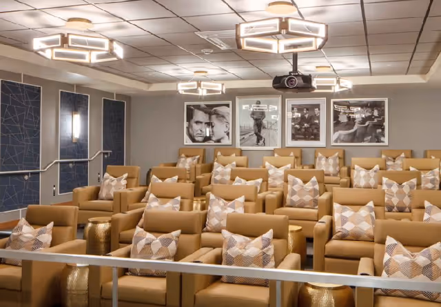 A modern theater room with multiple rows of tan leather recliner chairs, each with a patterned throw pillow. The room features geometric ceiling lights, framed black and white movie posters on the back wall, and a ceiling-mounted projector.