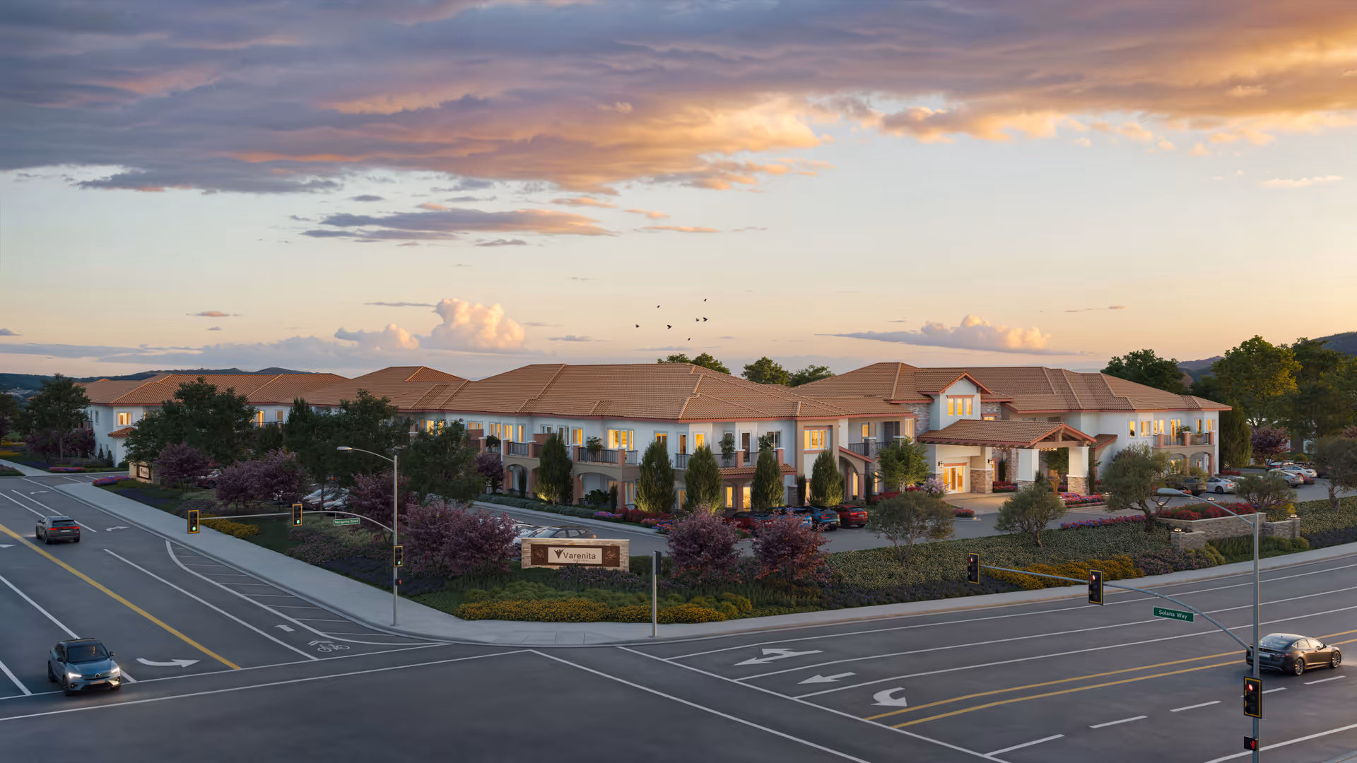 Exterior view of Varenita of Temecula, a large two-story senior living facility with a terracotta roof, surrounded by landscaped greenery and trees. The building is situated at a street intersection with traffic lights and cars on the road. The sky is partly cloudy with a warm sunset glow.