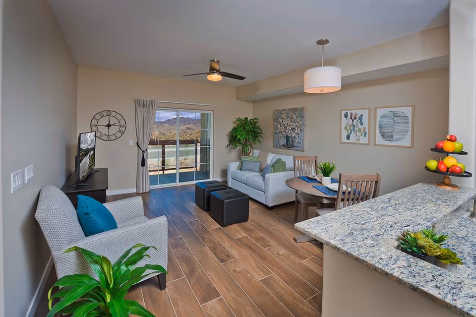 A bright and modern living room with wooden flooring, a gray armchair with a blue cushion, a white sofa with green cushions, two black ottomans, a round wooden dining table with two chairs, and a granite countertop with a tiered fruit stand. The room has a ceiling fan, pendant light, wall art, a large clock, and sliding glass doors leading to a balcony with a scenic mountain view.