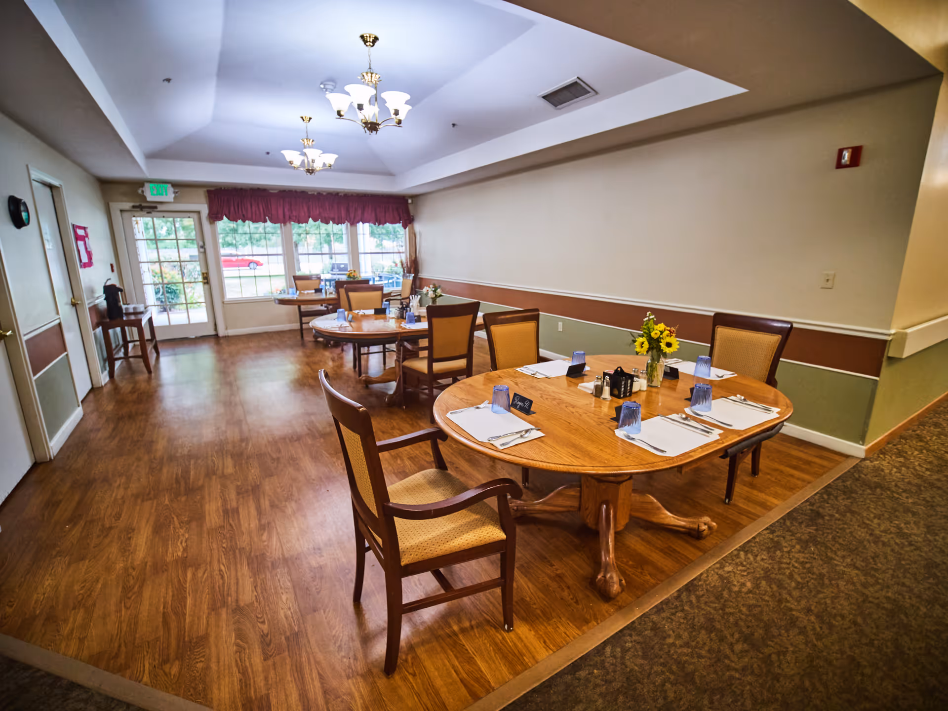 Dining area in Ascot Park Senior Living with round wooden tables set with placemats, utensils, blue drinking glasses, and small flower vases. The room has wood flooring, beige walls with a brown and green trim, and large windows with a red valance letting in natural light. There are chandeliers hanging from the ceiling and an exit door at the far end.