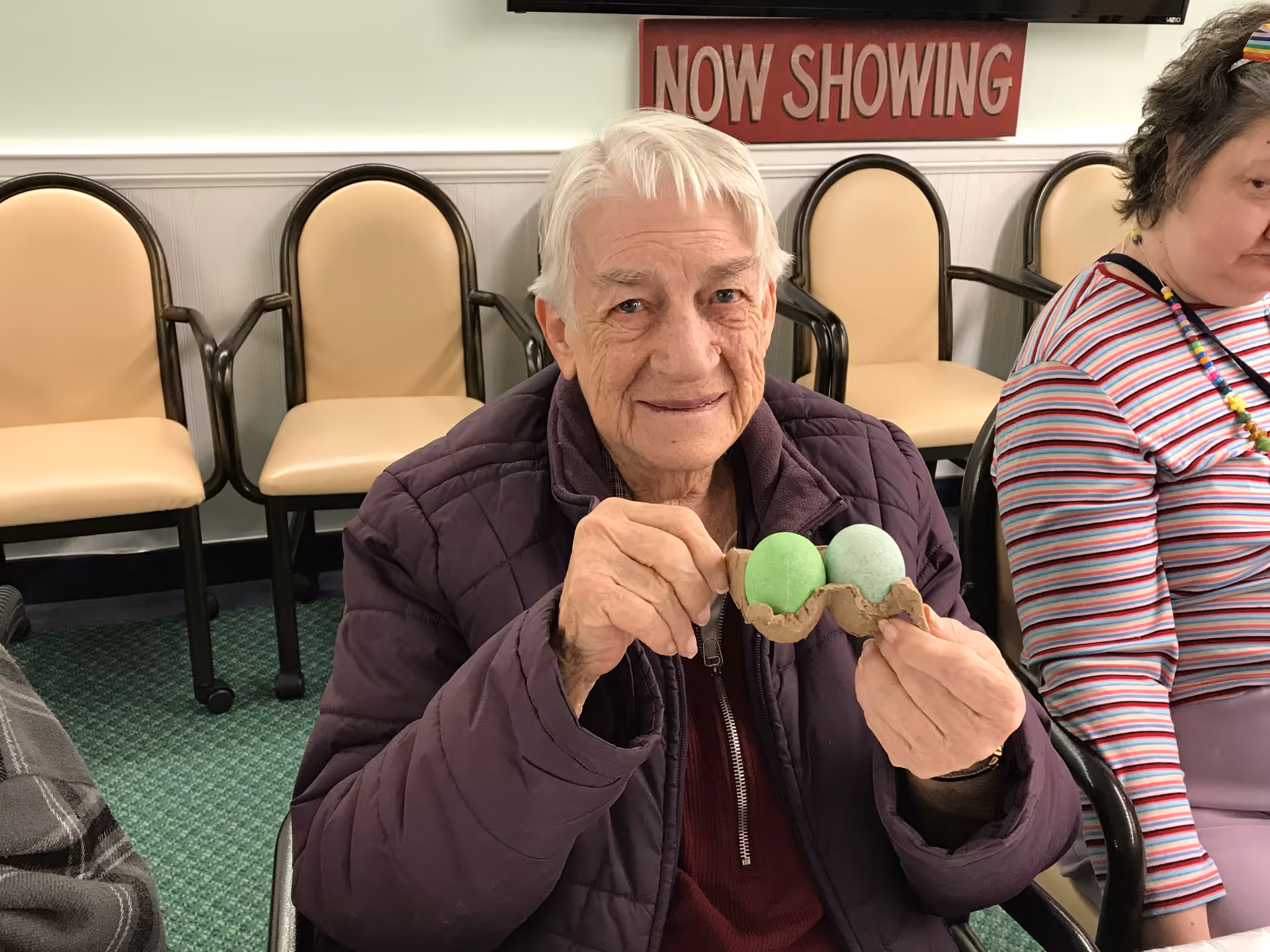 An elderly person with white hair wearing a purple jacket is sitting indoors and holding two green dyed eggs in a small carton. Behind them are beige chairs with dark frames and a red sign on the wall that reads 'NOW SHOWING'. Another person wearing a striped shirt is partially visible on the right.