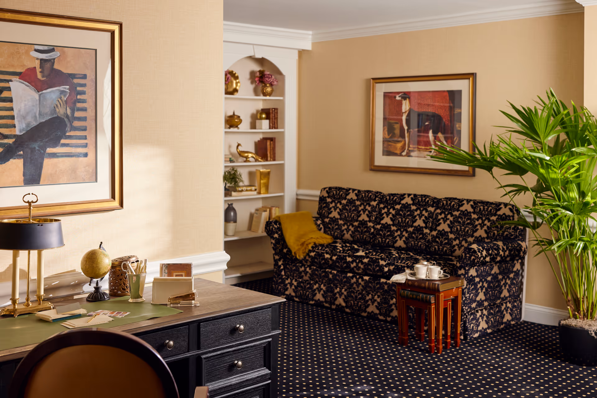 A cozy interior room featuring a dark patterned sofa with a yellow throw blanket, a small nested wooden table with a tea set, a large green potted plant, a black desk with various office supplies including a globe and lamp, and framed artwork on beige walls.