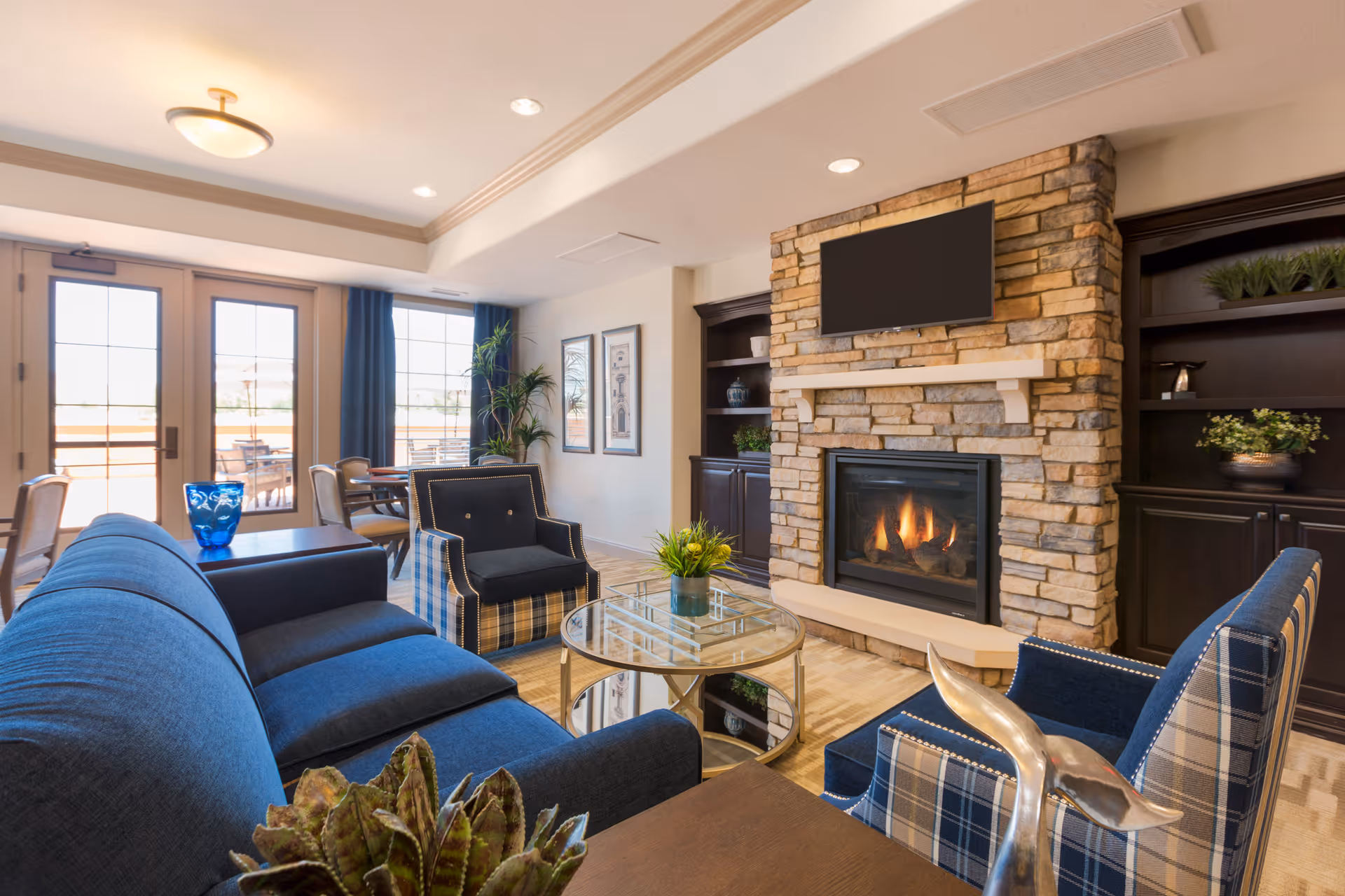 A cozy senior living room with a stone fireplace, a mounted flat-screen TV above it, and built-in dark wood shelves on either side. The room features a blue sofa, two plaid armchairs, a glass coffee table with a small plant, and a dining area with chairs and a table near large windows and a glass door leading outside.
