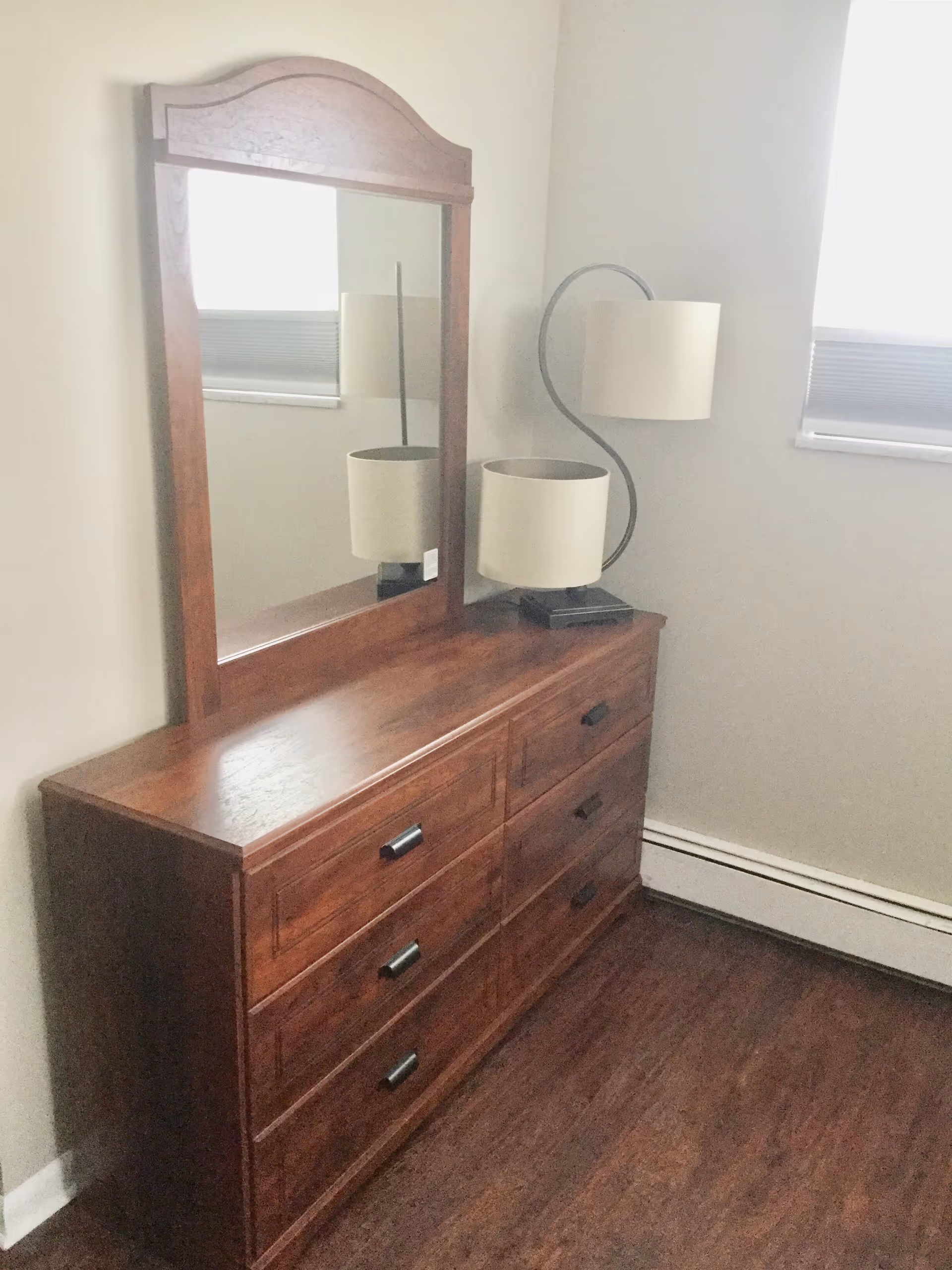 A wooden dresser with six drawers and a large attached mirror. On top of the dresser is a modern lamp with a curved metal stand and a white lampshade. The dresser is placed against a light-colored wall with a window nearby, and the floor is dark wood.
