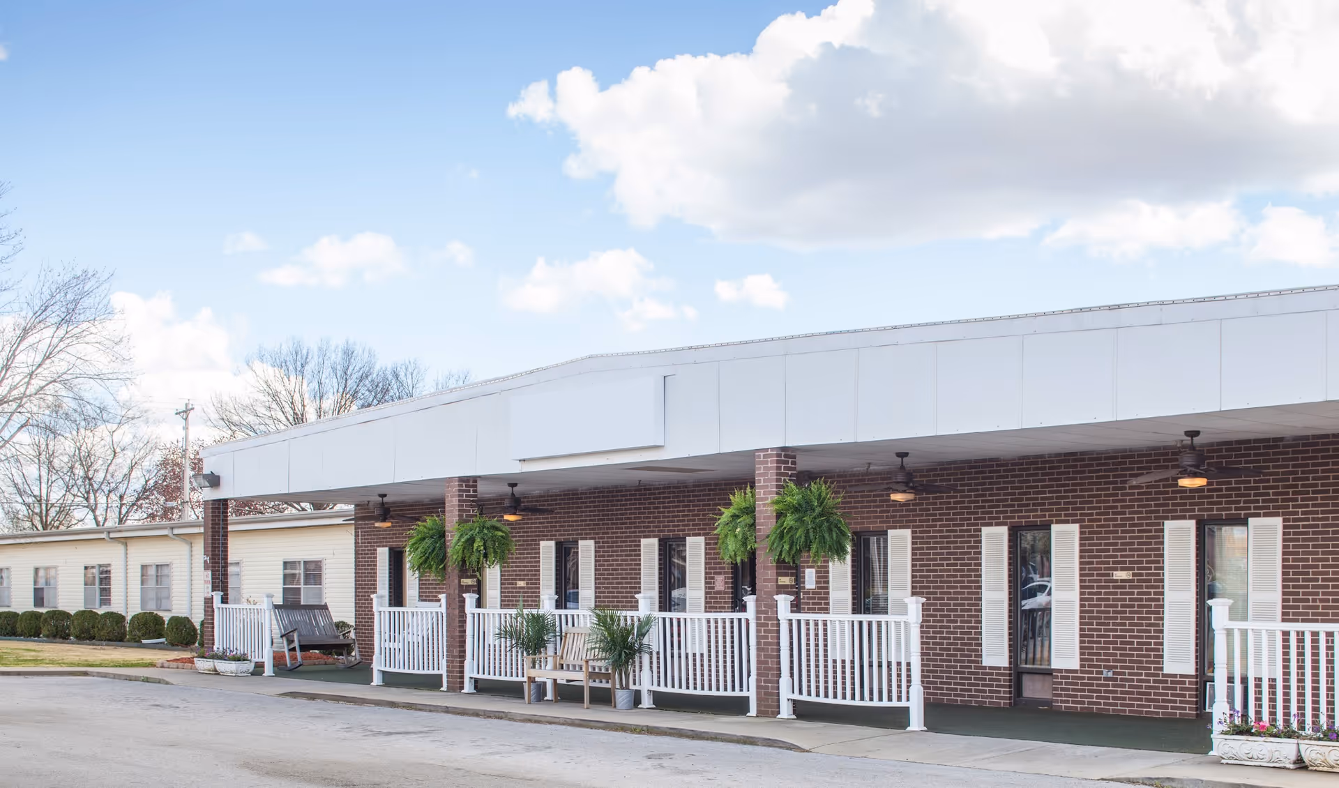 Exterior view of a single-story brick building with a covered porch featuring white railings, hanging green plants, and ceiling fans. The sky is partly cloudy and there are leafless trees in the background.