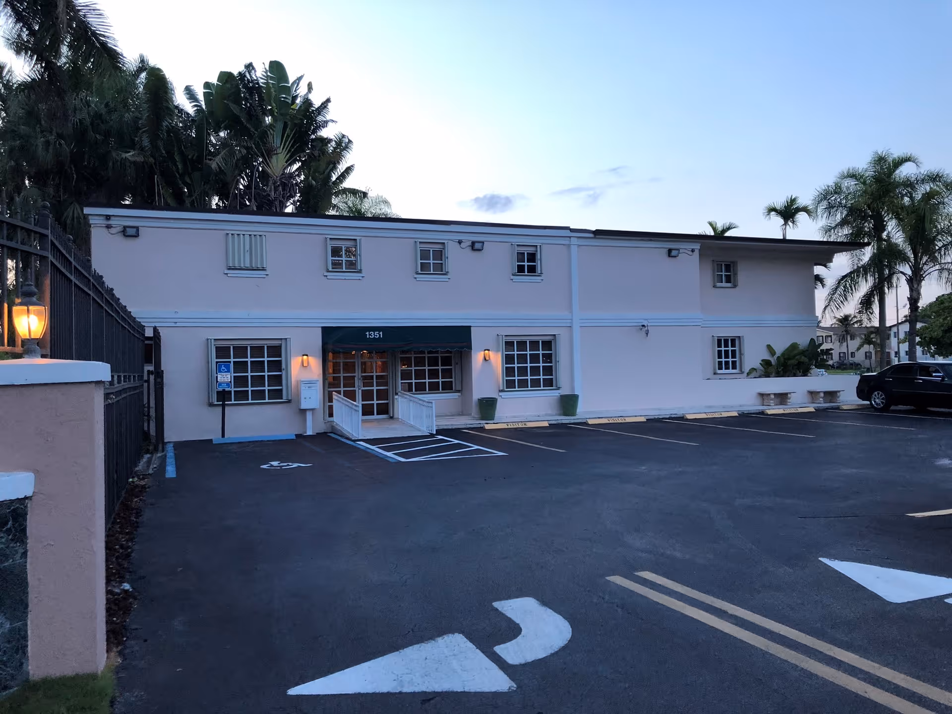 Two-story pale pink building front with a parking lot, handicap ramp entrance and palm trees in the background at dusk.