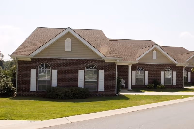 Exterior view of a single-story brick building with multiple arched windows with white shutters, a brown shingled roof, and a well-maintained green lawn in front. The building has a covered entrance supported by white columns.