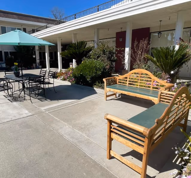 Outdoor patio area with two wooden benches with green cushions, a black metal table with four chairs, and a large green umbrella providing shade. The patio is surrounded by plants and flowers, with a building featuring white pillars and red doors in the background under a clear blue sky.