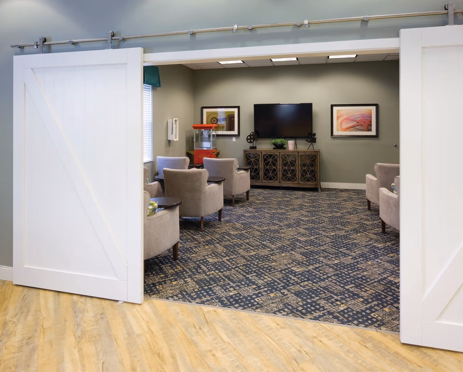 A cozy common area with beige armchairs arranged in a semi-circle facing a flat-screen TV mounted on a green wall. Below the TV is a decorative wooden cabinet with a plant and vintage film reel decor. There is a popcorn machine in the corner near a window with blinds. The room is accessed through white sliding barn doors and has a patterned carpet and wood flooring at the entrance.