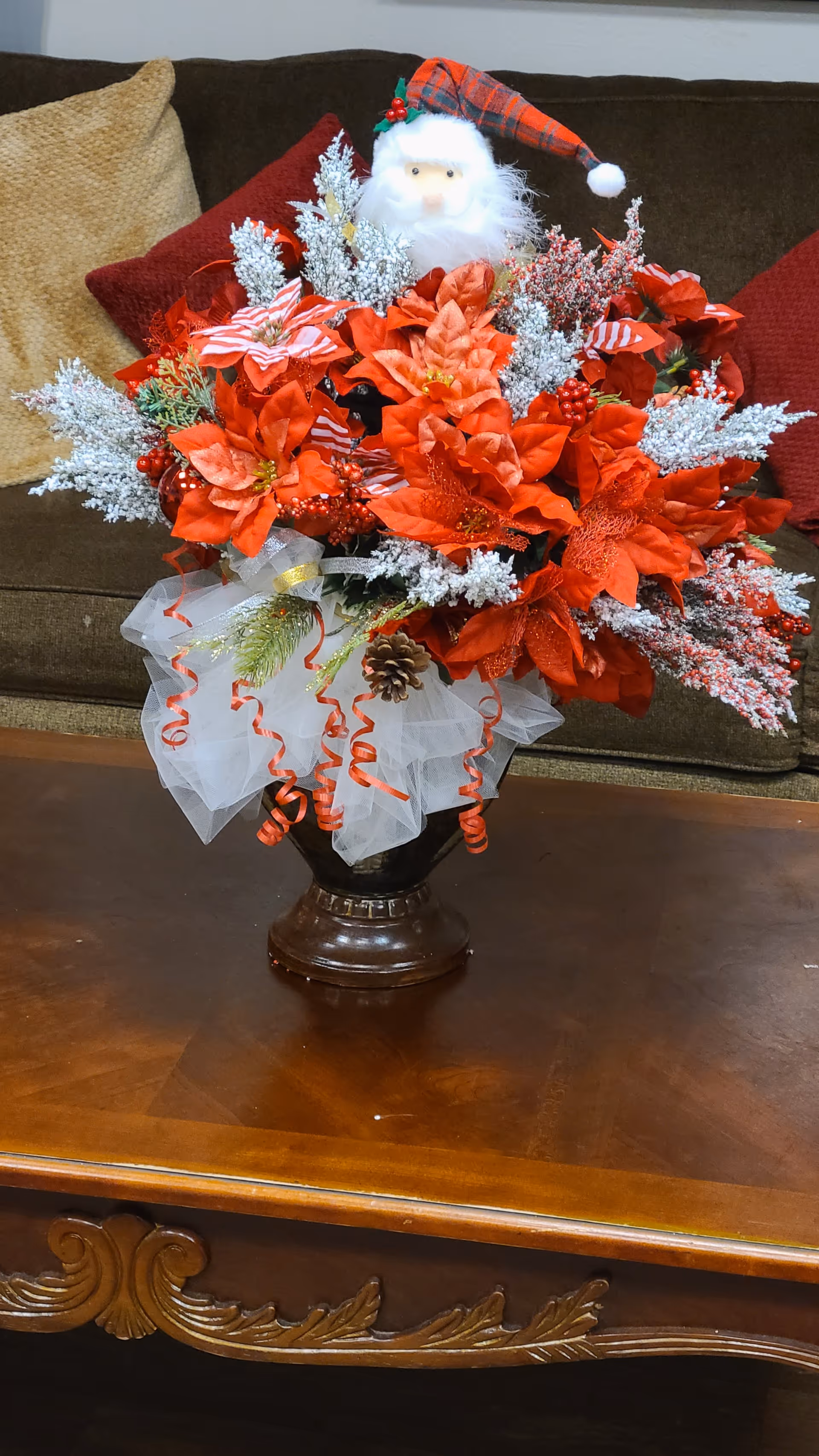 A festive arrangement of red poinsettias, white frosted branches and a small Santa figurine in a vase on a wooden coffee table with a couch behind it.