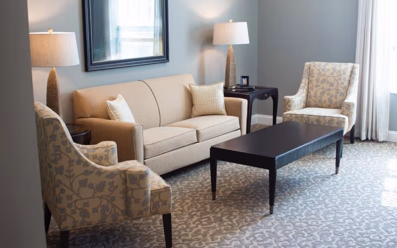 A well-lit living room with a beige sofa, two patterned armchairs, a dark wooden coffee table, and lamps on side tables.