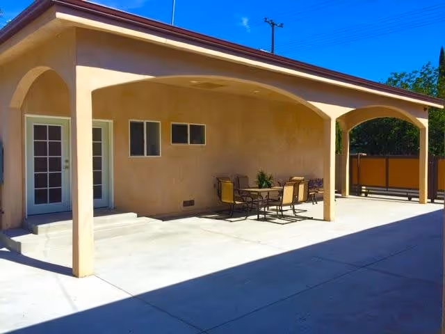 Covered exterior patio with arched openings, a table and chairs, and a concrete courtyard beside a beige building.
