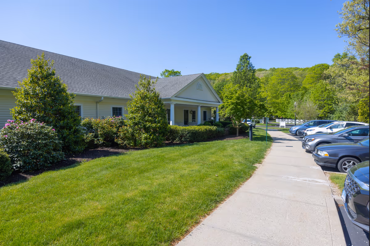 Exterior view of The Adel Center building with a well-maintained lawn, bushes, and trees under a clear blue sky. A sidewalk runs alongside the building leading to a parking area with several parked cars.