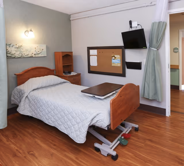 A single hospital-style bed with a gray quilted blanket in a room with wooden flooring. The bed has an adjustable overbed table on it. There is a wooden nightstand with shelves next to the bed, a wall-mounted flat screen TV, and a bulletin board on the wall. A green curtain is partially drawn to the side near the doorway.